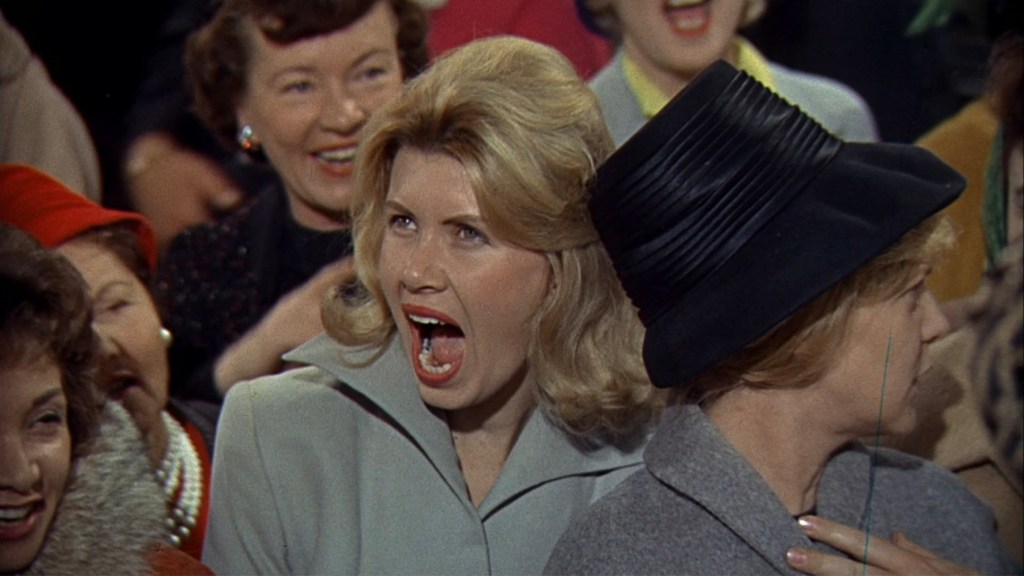 A blonde woman in a light coat passionately expressing excitement with her mouth open, surrounded by a crowd of smiling women, some wearing hats.