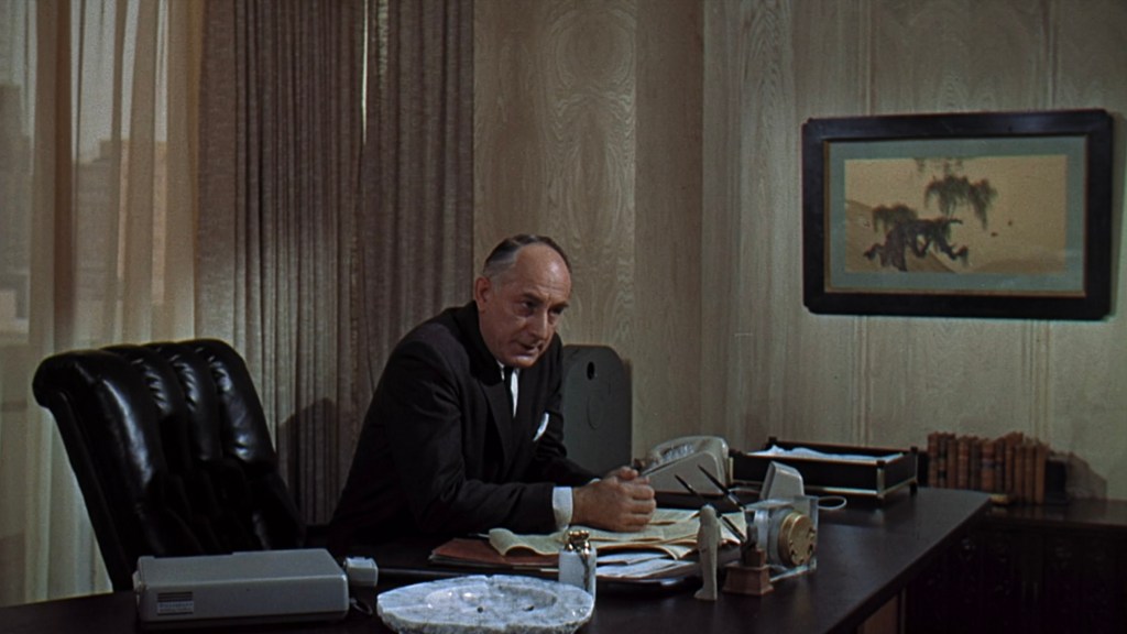 An older man in formal attire sits at a desk in a well-lit office, looking intently at paperwork. There are decorative elements, including a framed picture on the wall and office supplies on the desk.