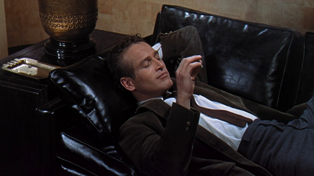 A man reclining on a black leather couch, appearing relaxed with a cigarette in hand, dressed in a jacket and tie.