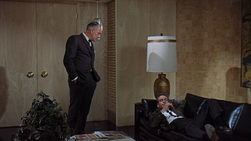 Two men in a modern interior; one is standing and looking down at the other, who is reclining on a black sofa with a cigarette in hand. A large lamp and decorative plant are visible in the background.