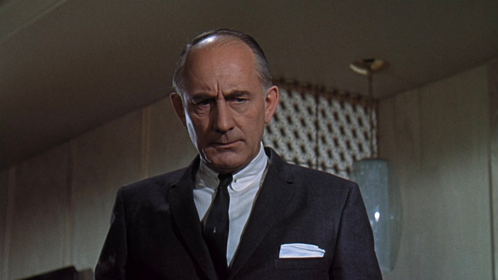 A serious-looking man dressed in a suit with a white pocket square, standing in an interior setting.