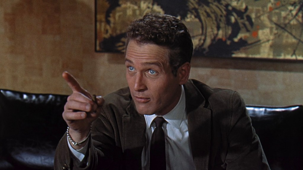 Man in a suit sitting on a sofa, pointing with his index finger and engaging in conversation.