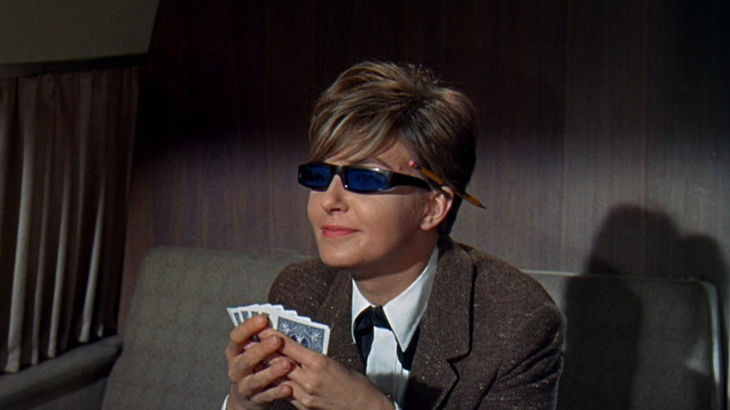 A woman wearing sunglasses and a suit, holding a hand of playing cards while sitting on a couch in a retro interior setting.