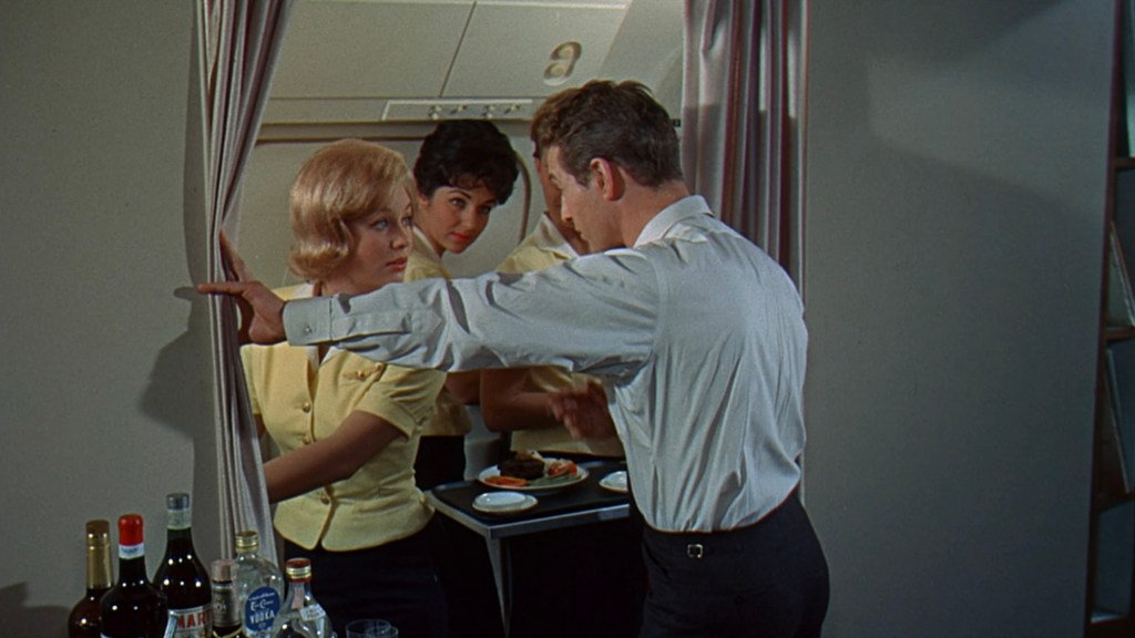 A scene set in an airplane interior featuring a man in a white shirt talking to a blonde woman in a yellow blouse while a dark-haired woman looks on. In the background, a small table displays various bottles and trays of food.