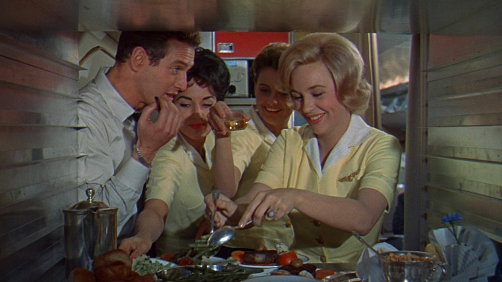 A scene inside an airplane kitchen showing a male flight attendant and three female attendants in yellow uniforms, sharing food and drinks while preparing a meal.