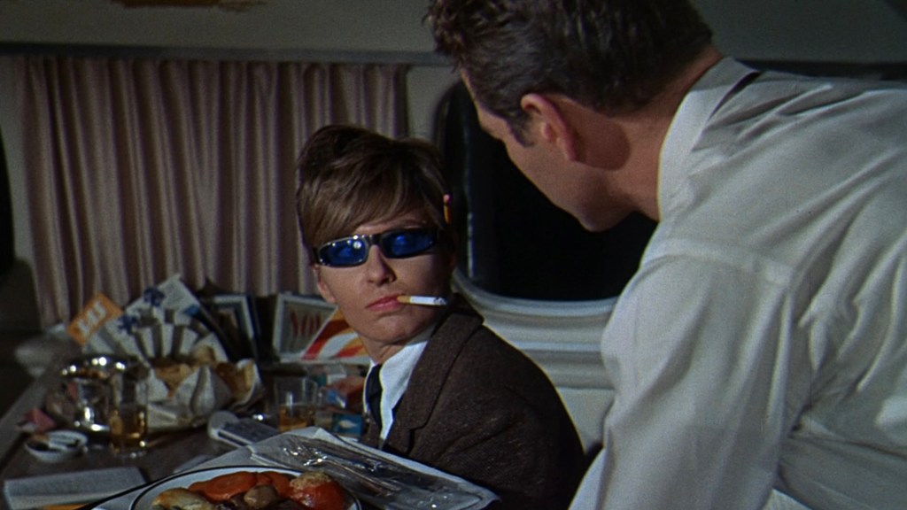 A character wearing sunglasses and a smoking cigarette looks intently at another person in a dining setting, with a table cluttered with food and drinks.