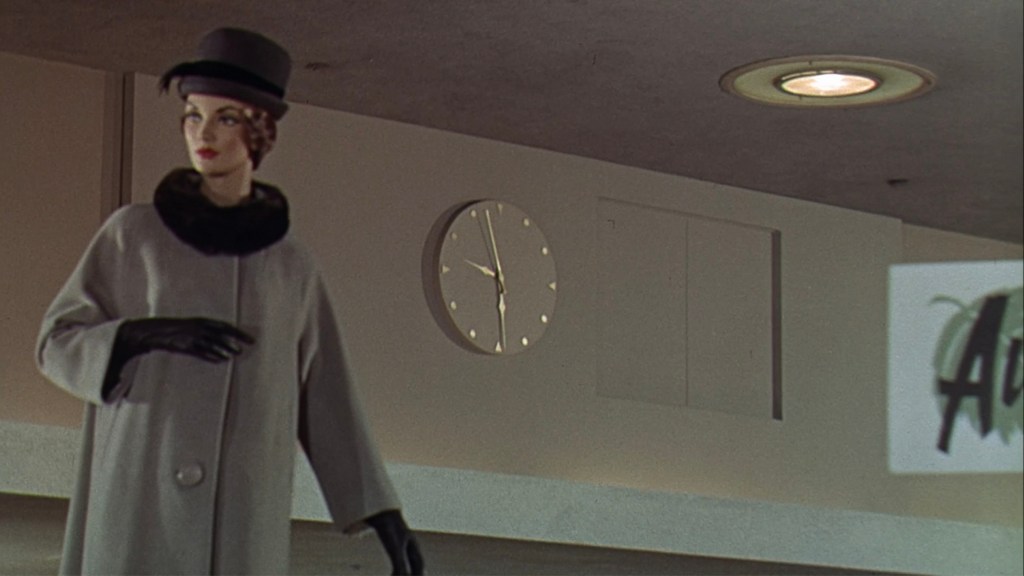 A mannequin wearing a grey coat and black hat, standing in a shopping environment, with a clock on the wall in the background.