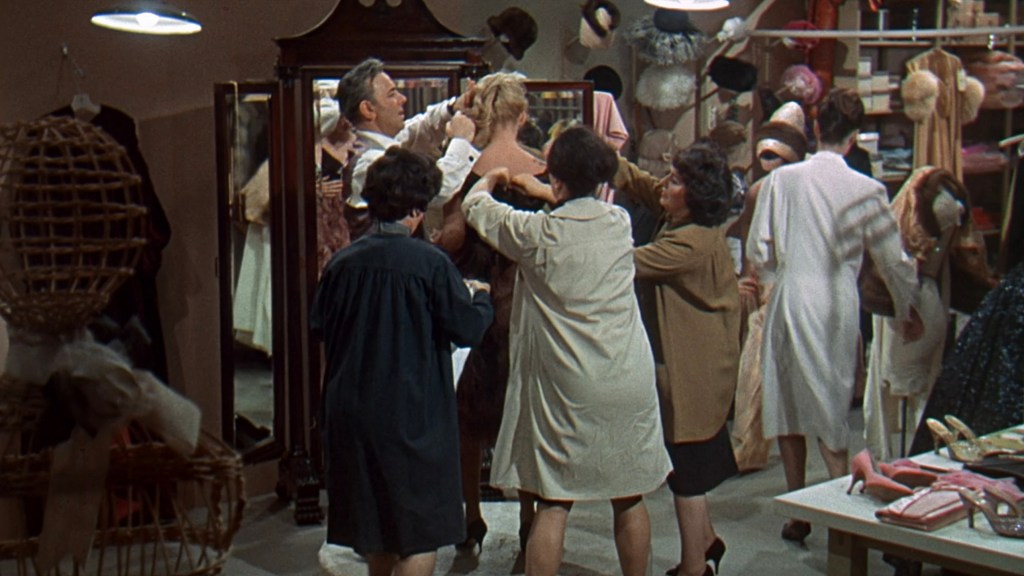 A chaotic scene in a clothing store where multiple women are fitting a dress on a model while a man adjusts it, surrounded by various fashion items and accessories.