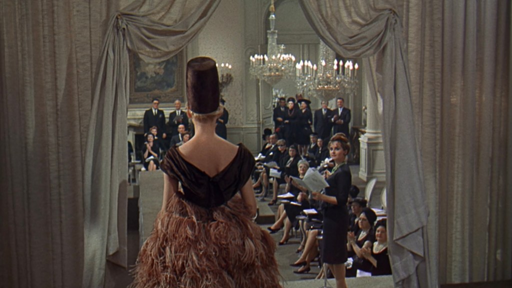 A model stands on a runway, wearing a dramatic feathered dress and a tall hat, while an audience dressed in formal attire watches intently. The setting features elegant chandeliers and decorative curtains.