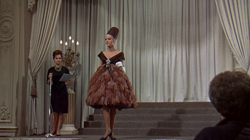 A fashion model walks the runway in a dramatic feathered dress while a presenter speaks from the side holding notes.