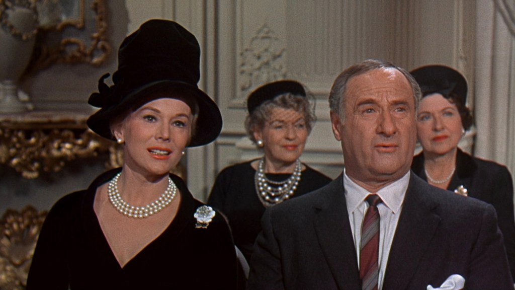 A woman in a black dress and large hat, adorned with pearls, speaks while a man in a suit listens intently. Behind them, three women in black hats observe the conversation, suggesting a formal or serious setting.