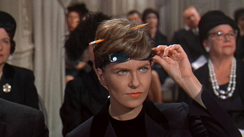 A woman with short hair styled up holds a headband with glasses on her forehead, appearing focused in a formal setting with other guests in the background.