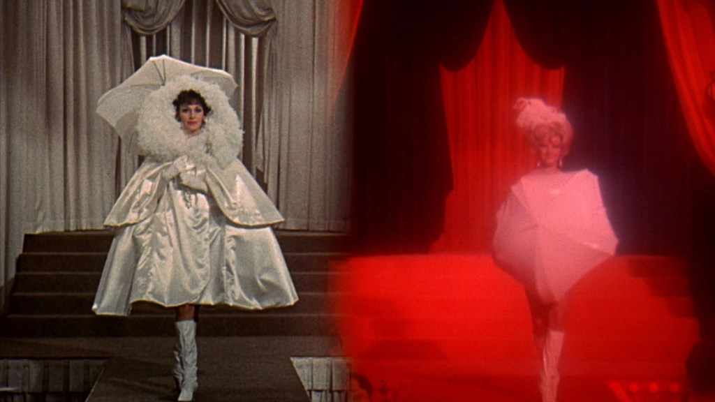 Two models on a runway, one wearing a large white dress with a fluffy collar and an umbrella hat, the other in a bright red setting with a white geometric outfit and headpiece.