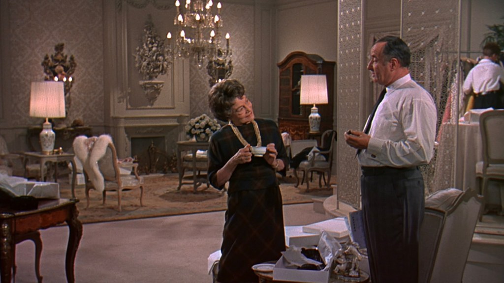 A woman in a black dress and pearl necklace stands in a lavish room, engaging in conversation with a man in a white shirt and tie. The opulent decor includes a chandelier, elegant furniture, and a cozy seating area.