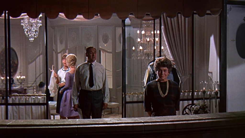 A captured moment showing four people in a stylish interior, with elegant lighting and decor. Two individuals are engaged in conversation while a woman stands apart, looking contemplative. The setting appears to be a formal gathering.