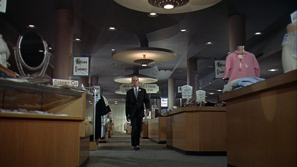 A man in a suit walking through a spacious department store aisle with display cases and clothing racks, featuring clearance sale signs and decorative lighting.