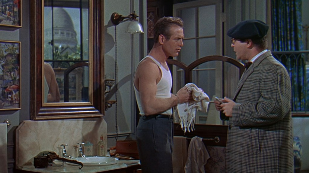 A scene in a bathroom featuring two men in dialogue. One man, wearing a tank top and pants, stands with a towel in his hands, while the other man, dressed in a checkered coat and hat, appears to be attentively listening. Reflections in the mirror and decorative elements are visible in the background.