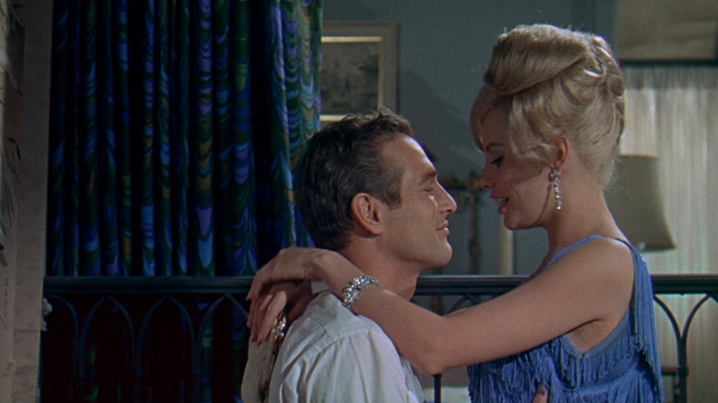 A man and a woman share an intimate moment, gazing into each other's eyes. The woman is wearing a blue fringe dress and has styled blonde hair, while the man has short hair and a light shirt. The background features colorful curtains and soft lighting.