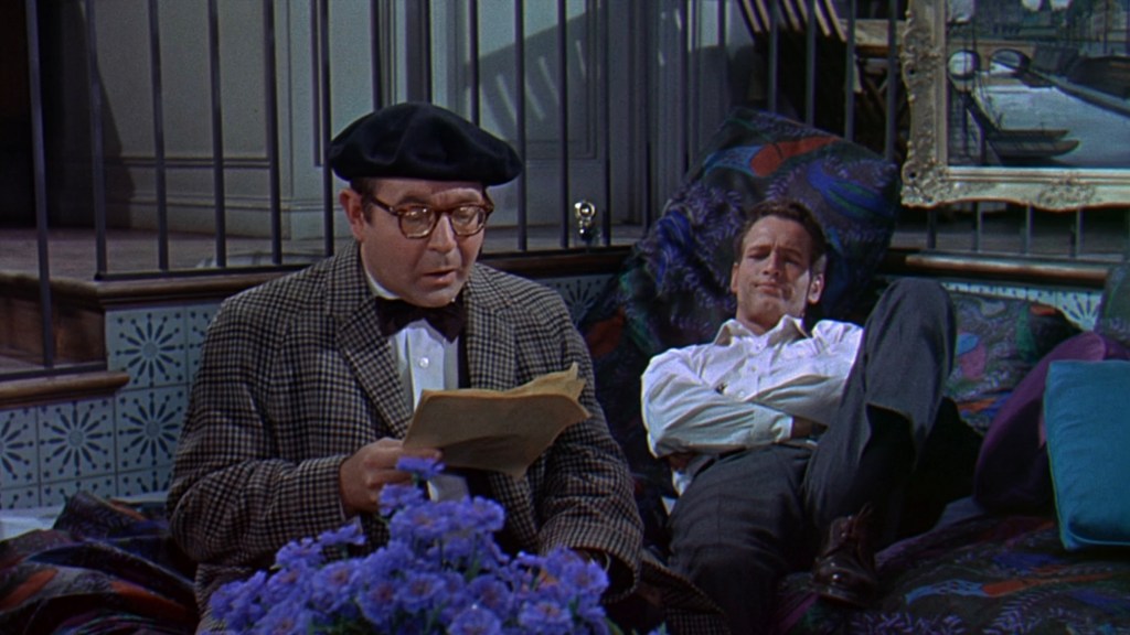 Two men seated in a colorful room; one reads a paper while the other reclines with a relaxed expression.