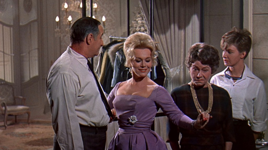 A scene featuring four individuals in a stylish interior, with one woman in a purple dress showing off a piece of jewelry while the others look on attentively.