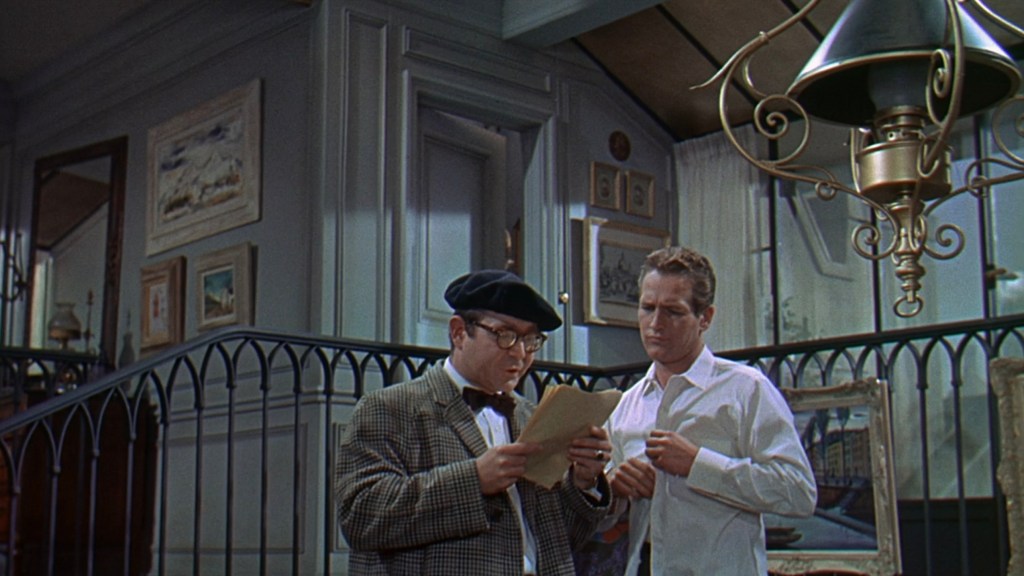 Two men in a well-decorated interior, one in a beret and glasses reading a document, while the other in a white shirt appears puzzled.