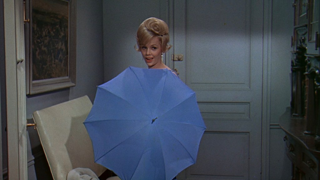 A woman with blonde hair holds a light blue umbrella, smiling while standing in a room with muted walls and simple furnishings.