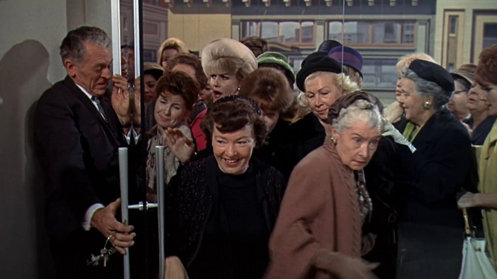 A group of women eagerly entering a building, with a man holding the door open. The women display a range of hairstyles and fashions from the era, indicating a lively atmosphere.