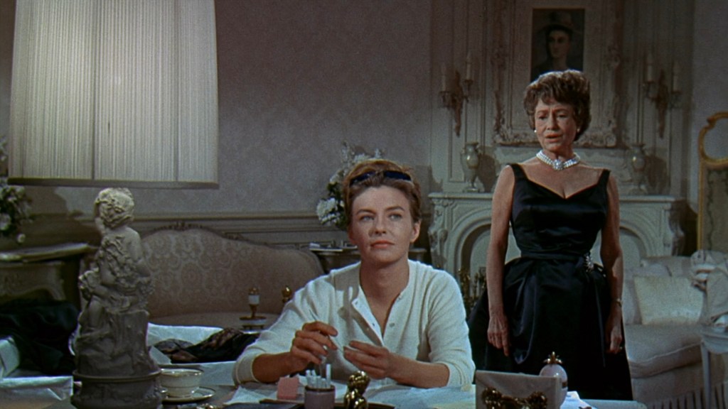 A woman in a white sweater sitting at a table with a beauty product in her hand, looking contemplative, while another woman in a black dress and pearl necklace stands nearby, observing her.