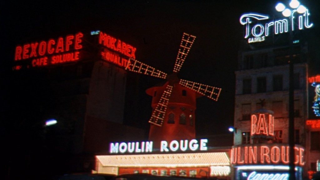 Nighttime view of the Moulin Rouge cabaret in Paris, featuring its iconic red windmill and illuminated neon signs including 'Rexocafé' and 'Formfit'.