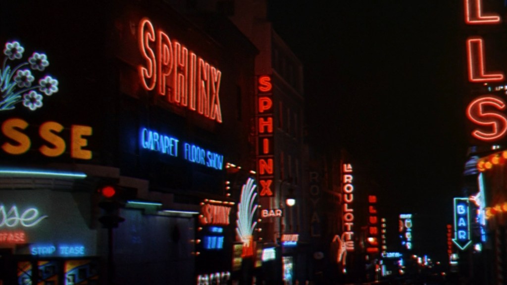 Neon signs illuminating a city street at night, featuring the names 'Sphinx', 'La Roulotte', and other establishments.