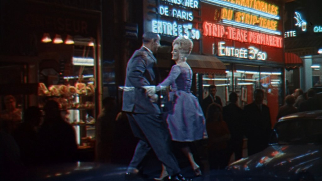 A couple dancing in front of a brightly lit strip club with neon signs showcasing entertainment options in a bustling urban setting.