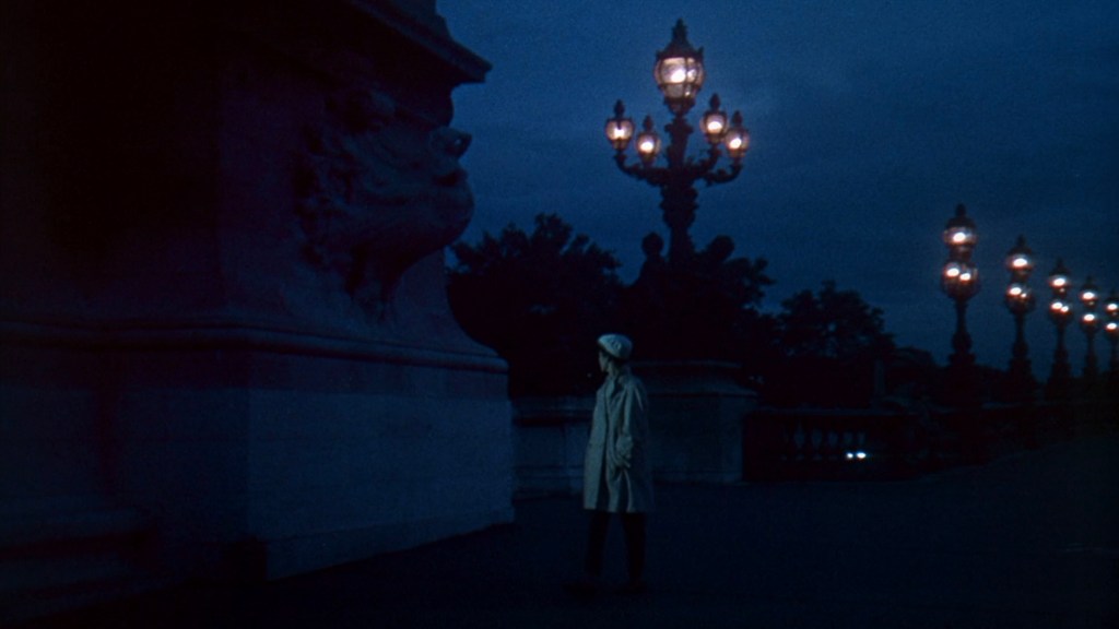 A figure in a trench coat stands on a dimly lit bridge at night, illuminated by streetlights, with a lion sculpture in the foreground and dark trees in the background.