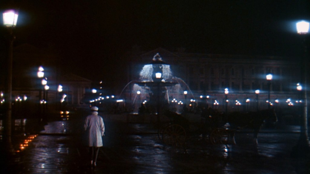 A nighttime scene featuring a fountain illuminated by street lamps, with a person in a white coat walking along a wet street, and a horse-drawn carriage in the background.