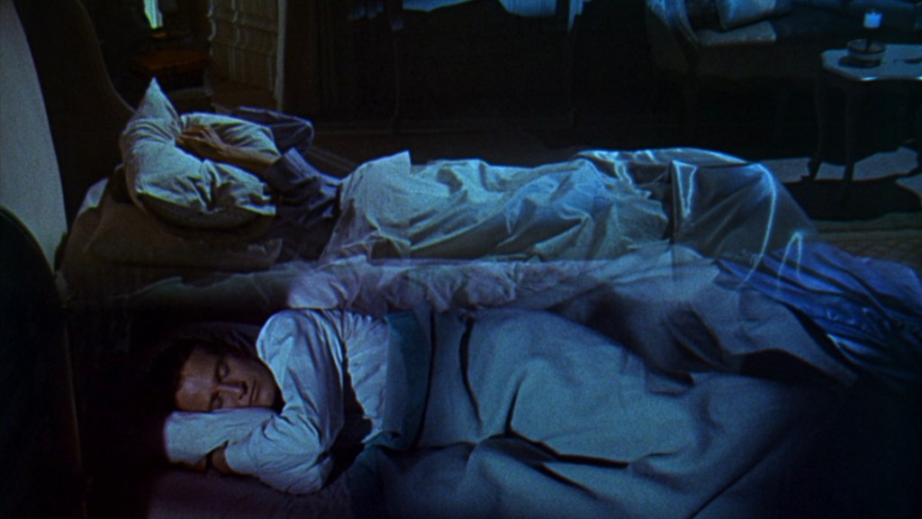 A dimly lit bedroom scene featuring two individuals sleeping in separate beds, each covered with blankets. One person is visible in the foreground, resting on their side with their head on a pillow, while the other is partially obscured in the background.