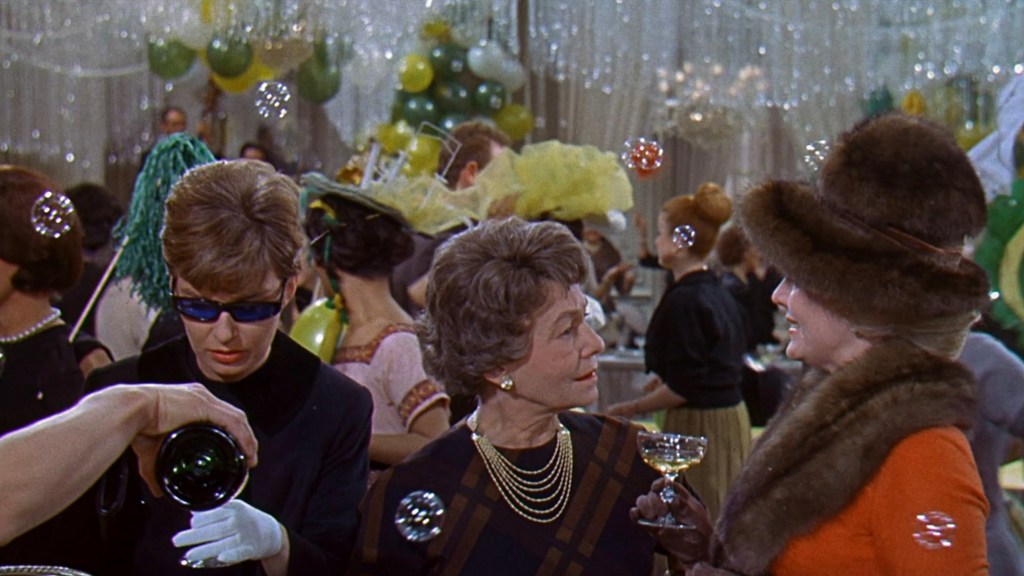 A scene from a festive party featuring three women in elegant attire, two of whom are engaged in conversation while one checks a drink. The background is filled with bubbles and party decorations, creating a lively atmosphere.