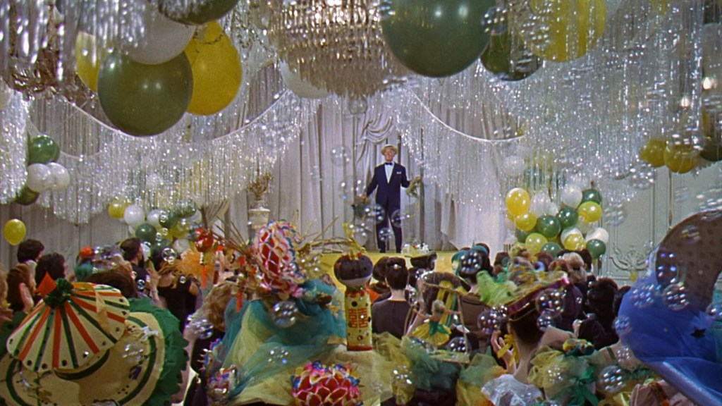 A vibrant party scene featuring colorful balloons and decorations, with a performer on stage addressing an audience dressed in festive attire and surrounded by bubbles.