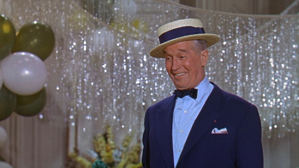 A smiling man wearing a straw hat and a tuxedo, standing in front of a festive backdrop with balloons and sparkling decorations.