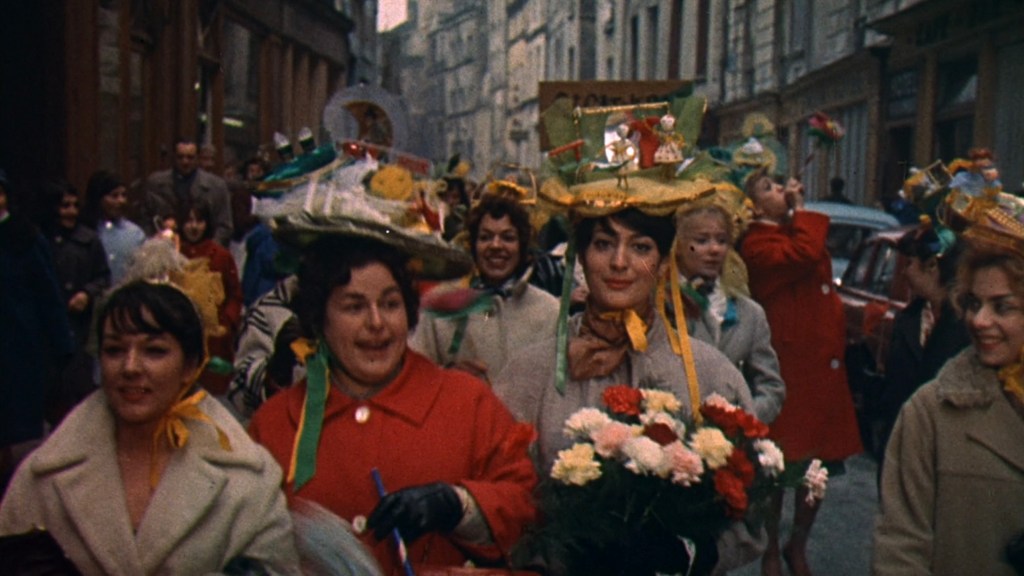 A lively street scene featuring a group of women wearing decorative hats and colorful outfits, some holding flowers. The women are walking in a parade-like fashion, with buildings and a car visible in the background.