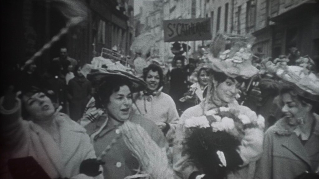 A black-and-white photo of a lively street parade with women wearing large decorative hats, celebrating with flowers and banners in a historic urban setting.