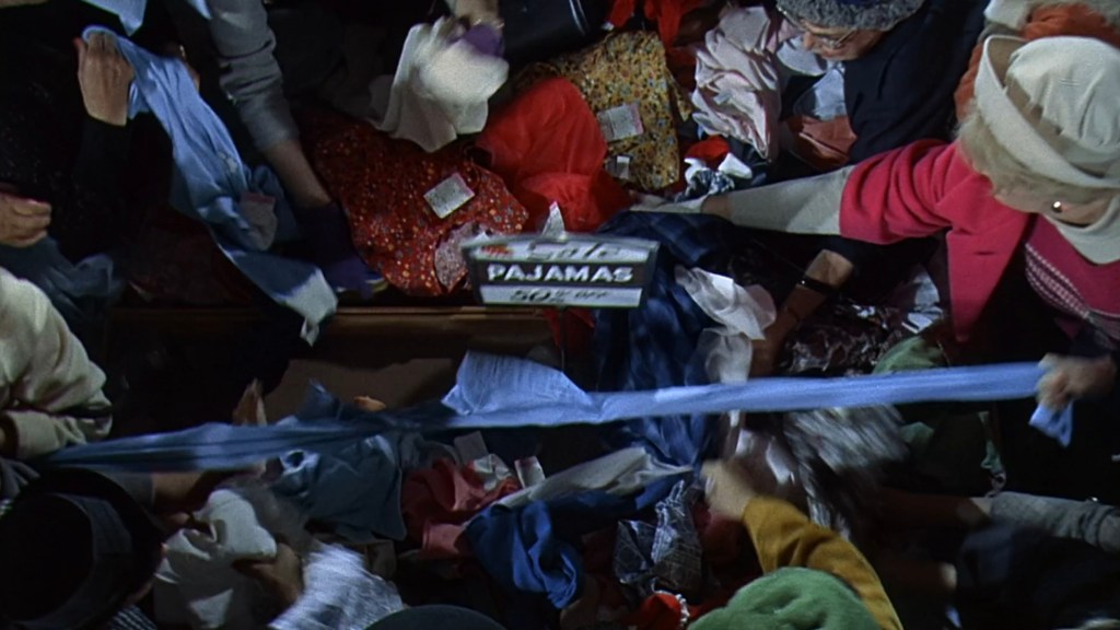 A chaotic scene of people rummaging through a pile of colorful clothes, with a sign labeled 'PAJAMAS' visible among the fabric.