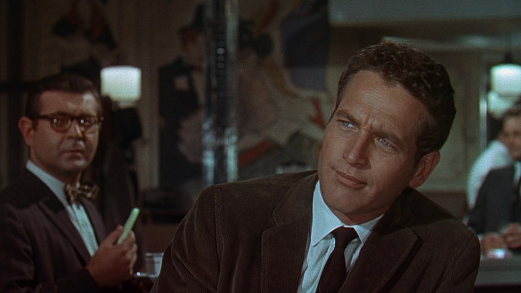 A scene featuring two men in a retro setting, one with curly hair and a suit, looking thoughtfully, while the other, wearing glasses and a bow tie, appears to listen intently.