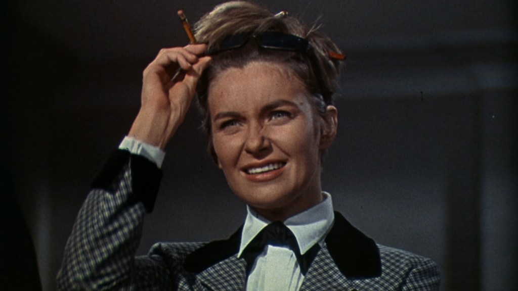 A woman with short hair and a puzzled expression, wearing a checkered suit with a black collar, holding a pencil in her hand and sunglasses on her head.