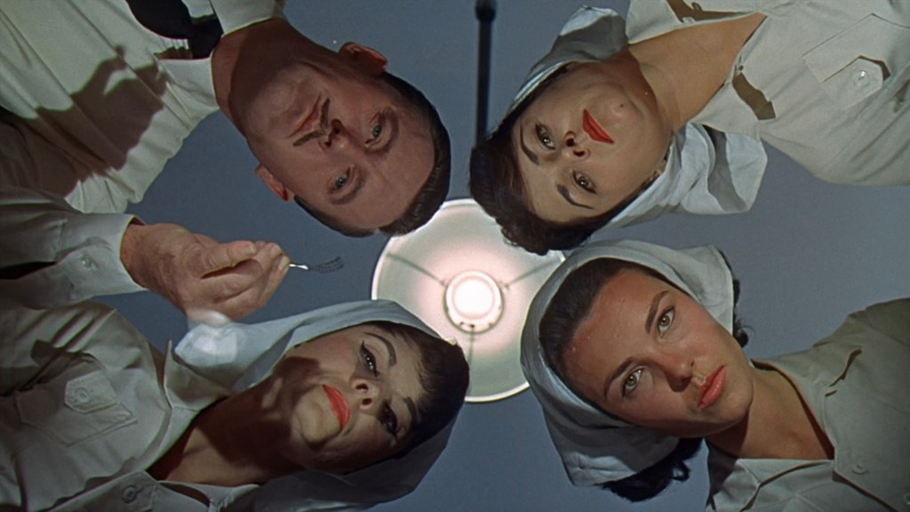 A group of four people, including two women in nurse uniforms and a man, are leaning over, looking down at the viewer. They are positioned around a bright overhead light, with expressions that convey curiosity or concern.