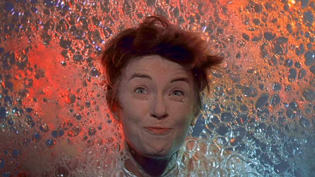 A person with short hair smiling playfully while surrounded by colorful bubbles and foam, creating a whimsical and vibrant atmosphere.