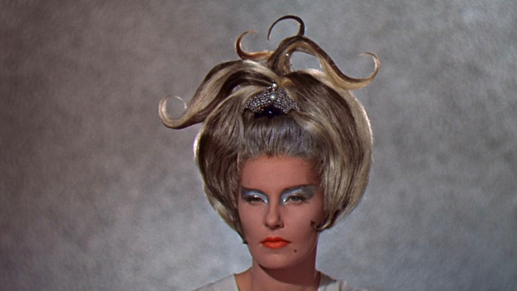 A woman with a dramatic, high hairstyle featuring spiraled curls, wearing bold blue eye makeup and bright orange lipstick, against a subtle textured background.