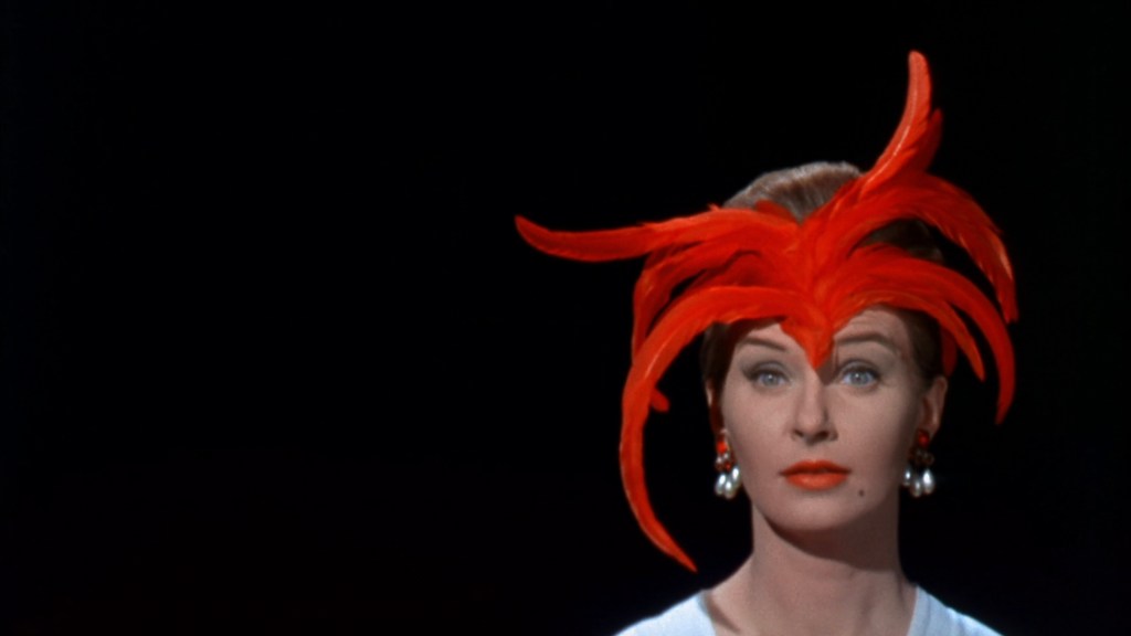 A woman wearing a dramatic red feathered hat and pearl earrings, with a neutral expression, against a dark background.