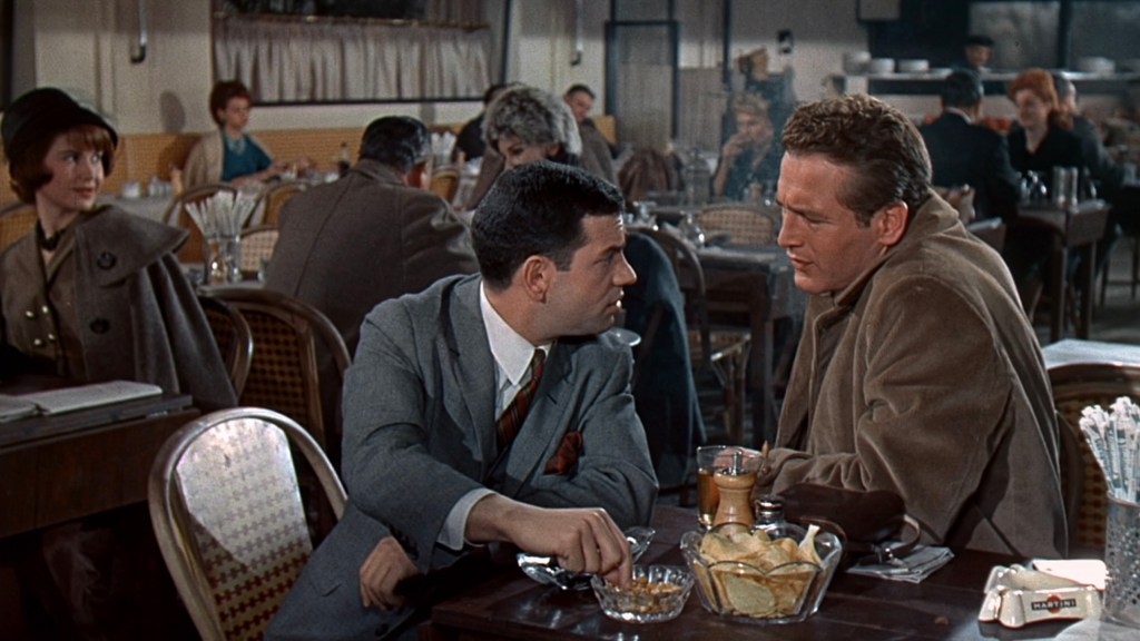 Two men sitting at a table in a restaurant, engaged in conversation. One man is dressed in a gray suit, while the other is wearing a brown coat. The background features other diners and typical restaurant decor.