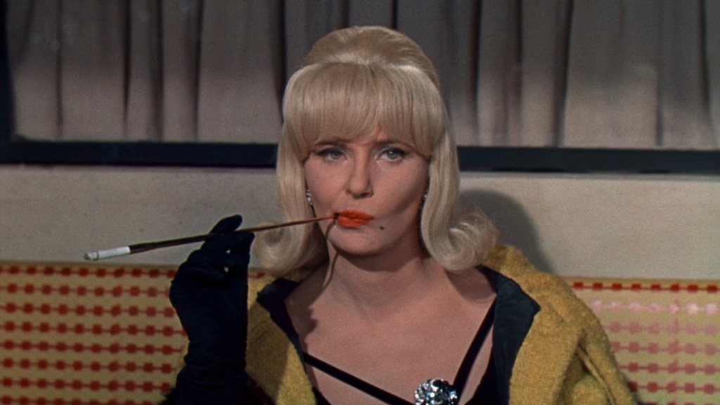 A woman with blonde hair styled in waves, wearing a black dress and yellow shawl, sits against a patterned background. She has a thoughtful expression while holding a cigarette holder with red lipstick on her lips.