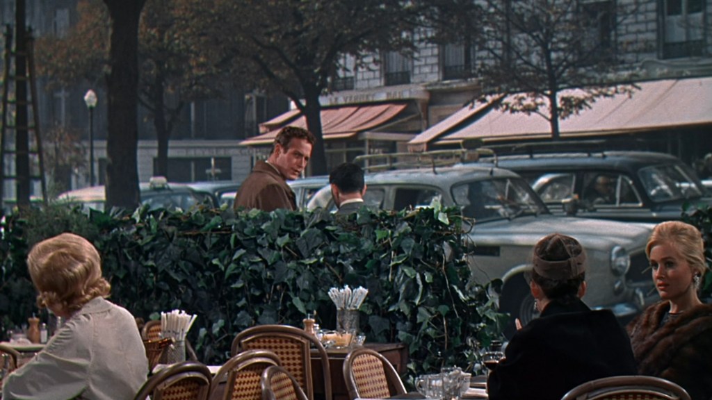 A street café scene featuring several diners sitting at tables, with a view of pedestrians and cars in the background, framed by green foliage.