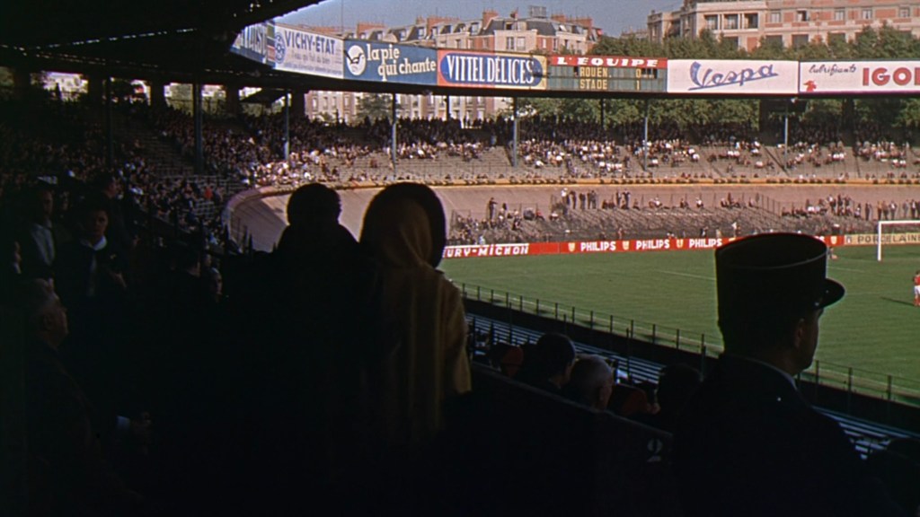 A view of a crowded stadium with spectators seated in the stands, banners and advertisements visible above, and a green field in the background.